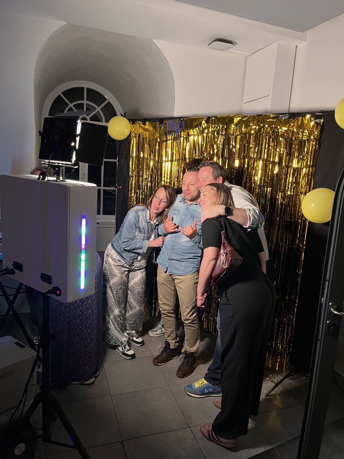 Quatre amis prennent la pose devant le photomaton Photoboox, rideau doré et ballons jaunes